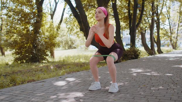 Athletic fit slim sport girl doing squats using fitness elastic rubber band as part of her fitness workout outdoor in park at morning. Young woman enjoying healthy lifestyle. Active sportswoman jogger