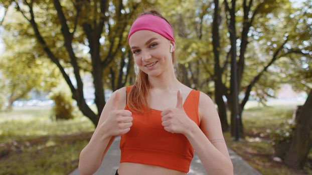 Portrait of athletic fit smiling runner girl training looking at camera, happy about marathon achievement. Workout cardio in park. Woman raises thumbs up agrees with something or gives positive reply