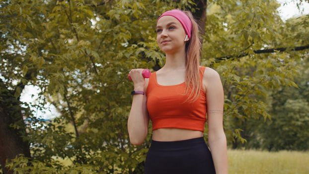 Athletic young sporty fit girl doing dumbbells workout in park performing bicep curls to build arm muscle and strength. Pretty woman enjoying exercising outdoors. Active sportswoman sport motivation