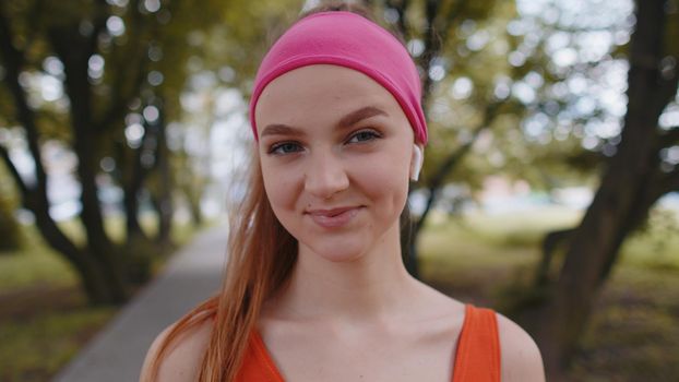 Athletic fitness sport runner adult girl training, listening favorite music song in earphones. Young woman looking at camera and smiling, laughing. Workout cardio in park. Active sportswoman jogger