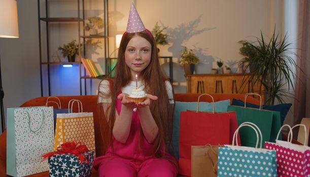 Happy teen child girl wears festive birthday hat hold cupcake makes wish joyful congratulating blowing burning candle on cake. Ginger kid celebrating anniversary party on couch with gifts at home