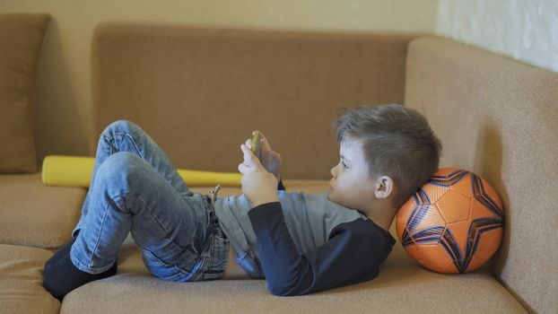 Young Boy Spends Time Playing Games On Mobile Phone At Home, Little Caucasian Guy In Casual Clothes Lies On Sofa Putting Ball Under His Head, Maybe He Has Dopamine Addiction, Quarantine Concept