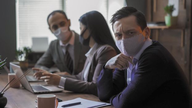 Business People In Protective Masks At Meeting In Office Despite Quarantine, Focus On Serious Young Man Communicates With Colleagues Sitting At Table In Board Room