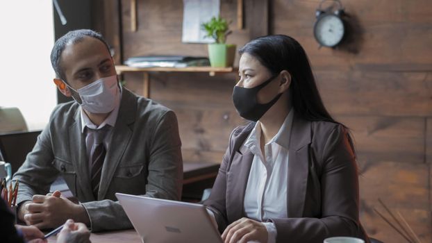 Business Team In Medical Masks Meetings In Modern Office During Quarantine, Man And Woman Talks Sitting At Desk, Entrepreneurs Keep Social Distance Due To Coronavirus Outbreak