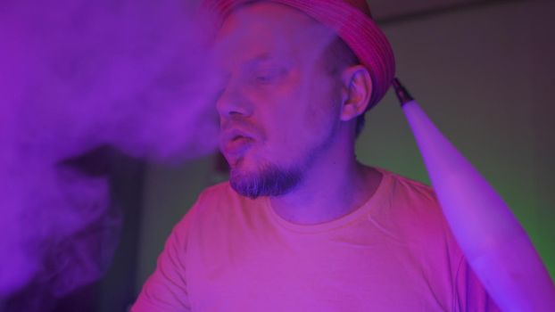 Smoker Of Hookah Exhales Smoke In Neon Lighting, Creative Man In Hat Smoking Hookah Sitting In Foggy Dark Room With Fluorescent Illumination, Quarantine Concept