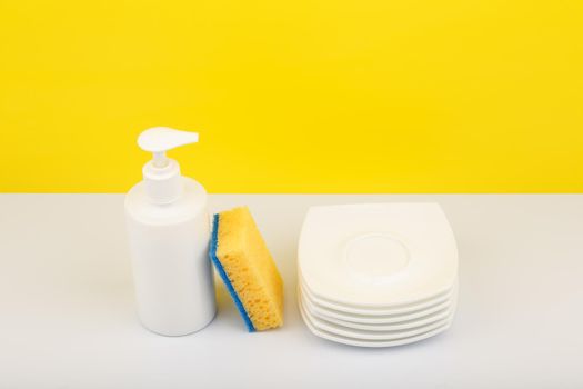 Dishwashing concept, high angle view of liquid detergent in white plastic bottle with dispenser, yellow cleaning sponge and pile of clean plates. Creative composition with copy space