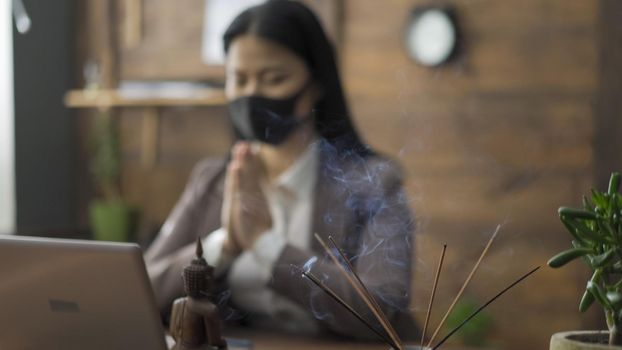 Prayer Of Asian Business Woman Sitting In Mask On Her Face At Office Table In Front Of Laptop, She Clasped Hands Together Reading Prayer, Business During The COVID 19 Pandemic