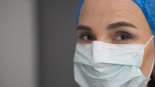 Doctor In Protective Mask And Blue Cap Looking At Camera, Female Medic Exhausted By Work During An Outbreak Of Corona Virus, Selective Focus On Right Eye, Close Up Of Doctor's Face, Three Quarter View