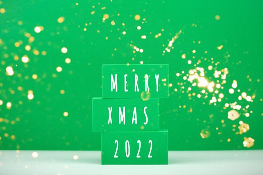 Merry Xmas 2022 concept in green colors with golden bokeh. Stack of toy blocks with text on white table against green background with firework. Creative minimal Christmas composition