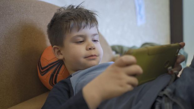 Child Looks At Phone, Little Boy Plays On Mobile Phone While Lying On Sofa, He Uses Ball Instead Of Pillow, The Boy Is Forced To Be Isolated Due To Coronavirus Epidemic, Quarantine Concept