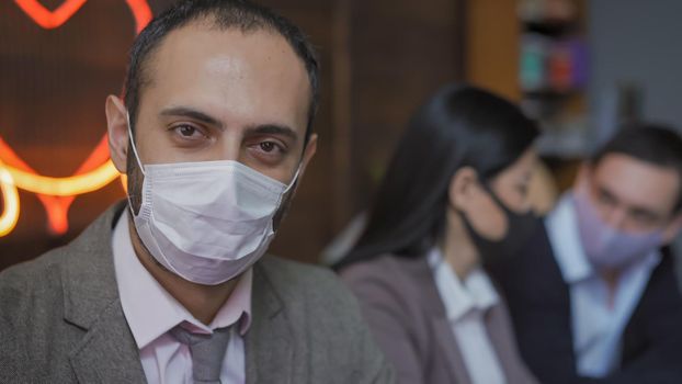 Attractive Middle Eastern Man In Mask Sitting At Business Meeting In Modern Office, His Colleagues Chatting On Blurry Background With Neon Lighting, Quarantine Concept