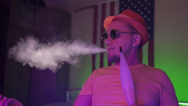 Man Exhales Smoke In Neon Backlit, Profile View Of Creative Man Wearing Sunglasses And Hat Smoking Hookah Against The Background Of American Flag, Quarantine Concept