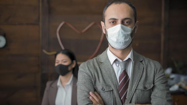 Business Man In Protective Mask Working During Epidemic, Man Looks At Camera With His Arms Crossed While Standing Against The Background Of Wooden Wall And His Female Colleague