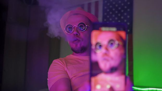 Man Making Selfie By Phone In Neon Lighting, Selective Focus On Hipster Wearing Sunglasses And Hat Exhales Smoke Smoking Hookah And Using Smartphone In Self Isolation, Quarantine Concept