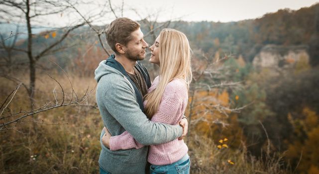 Lovely Couple Of Travelers Stands Embracing On Cliff Edge Outdoors, Smiling Man And Woman Are Hugging And Looking At Each Other While Standing Against Background Of Autumn Nature