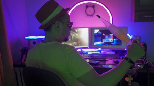 Man Smokes Hookah Working At Home During Quarantine, Man Sitting In Dark Room With Neon Lights And Smoking Hookah, Self Isolation Quarantine Days, Rear View