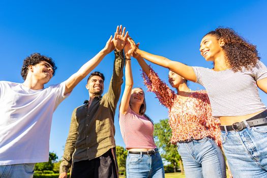 Group of happy gen z friends touching hand raised up to each other. Concept of trust in future and success in life. Diverse millenial multi ethnic students bonding outdoor in nature of city park