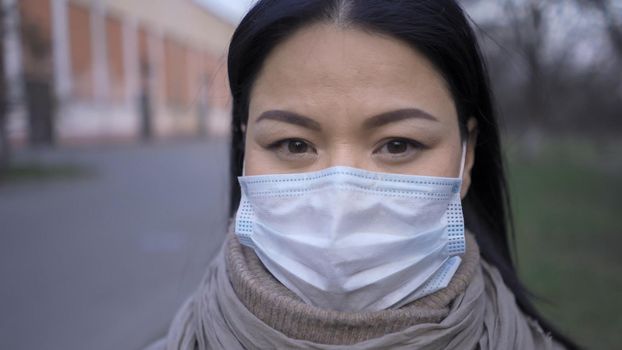 Asian Woman In Protective Mask Stands Outdoors, Beautiful Brunette Looks At Camera Standing Against The Background Of The Old Factory Building, Close Up Of Woman's Face, Quarantine Concept