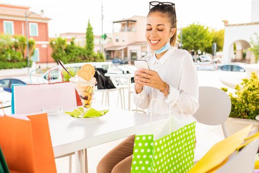 Happy young woman resting with open protective face mask sitting at a bar outdoor among several colored paper bag to eat ice cream after shopping in city center. Girl using smart phone to buy online