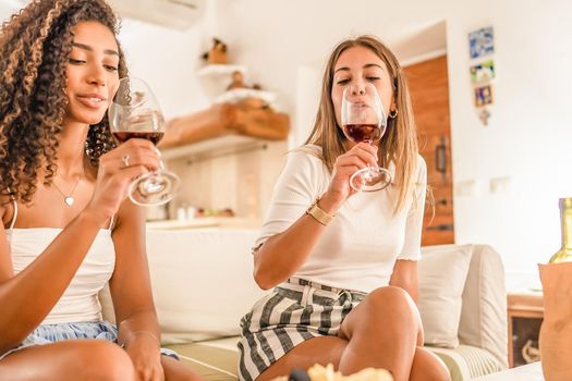 Two young women celebrating at home drinking red wine into glasses to toast happily. Caucasian gay girl bonding with her Afro-American curly girlfriend drinking on sofa. Concept of alcohol abuse