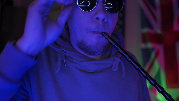 Hipsters Smoking Hookah Having Fun Self-Isolation Time At Home In Neon Backlight, British Man In Sweatshirt Corrects Sunglasses Looking At Camera Against The Background Of Great Britain Flag