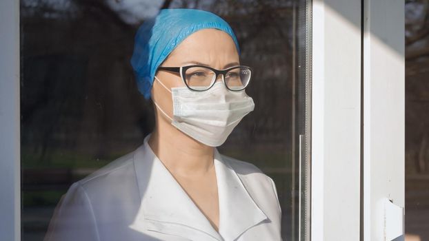 Female Medic Looks At Window Standing Behind Glass In Isolation On Sunny Day, Tired Woman In Protective Mask And Uniform Looks At Window Waiting For End Of Epidemic, Pandemic Concept