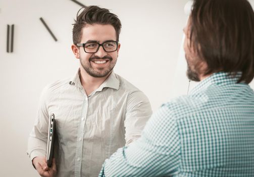 Greeting colleagues shaking hands, two employees greeting each other shaking hands, selective focus on a smiling man in glasses standing against a bright office wall. Toned image.