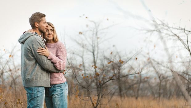 Loving couple resting while standing embracing on a background of autumn natural landscape. Young caucasian man and woman hugging on blurred young forest background. Copy space on the right side.