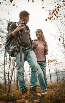 Tourists Backpackers Walking In Autumn Nature On Fallen Leaves And Green Grass, Focus On Man Holding His Woman's Hand And Hiking Poles In Other Hand, Walk With Backpacks Concept, Shot From Below