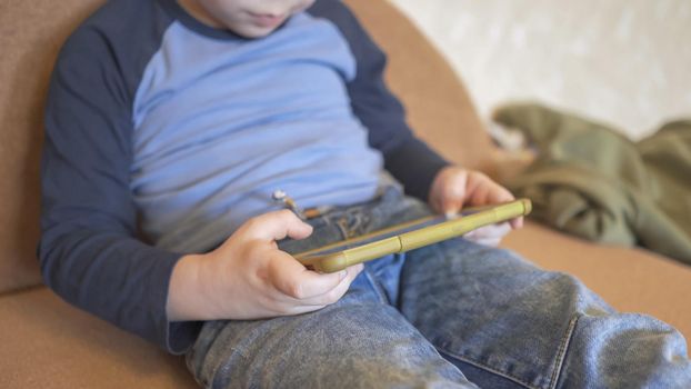 Boy Uses Phone Sitting On The Couch, Caucasian Boy In Casual Wear Spends Time Playing Or Learning Over Phone At Home, Little Boy Needs To Stay At Home Due To Quarantine, Close Up, High Angle View