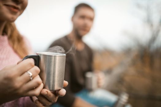 Two tourists drink a hot drink in nature. Close up of metal camping cup with steam from a hot coffee or tea in female hands. Caucasian guy with smiling girl have a rest outdoors. Close up shot.