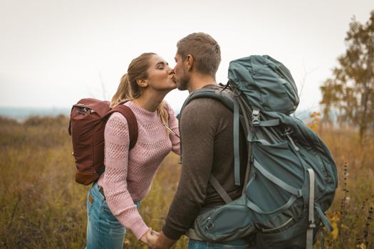 Loving backpackers kiss in the field outdoors. Happy couple of travelers, young man and woman standing against the backdrop of autumn nature. Love for adventure concept.