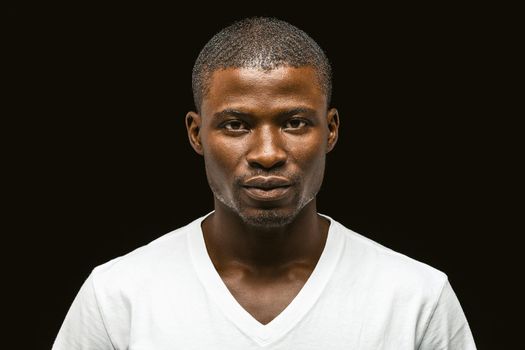 Serious African american man looking at the camera, attractive dark-skinned young guy in a white t-shirt posing expressing confidence isolated on black background. Toned image.