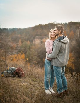 Young tourists stand embracing on the edge of a cliff inhaling the aromas of autumn nature, their backpacks are next to them on the grass