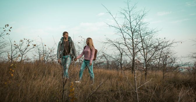 Tourists With Backpacks Are Hiking Together, Young Couple Of Travelers Is Walking On The Lawn Holding Hands, Cheerful Man And Woman Walking Outdors, Healthy Lifestyle Concept