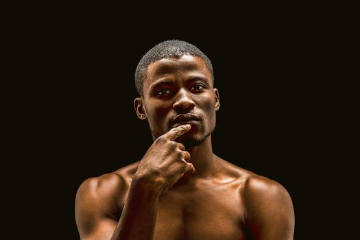 Naked African man thoughtfully looks at the camera while touching the fingers of his lips, sexy muscular young guy posing in the studio, cut out on a black background. Toned image.