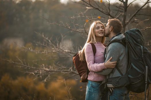 Men and women with backpacks stand gently embracing against the backdrop of autumn nature on the edge of a cliff. Young man kisses his beautiful blonde wife outdoors.