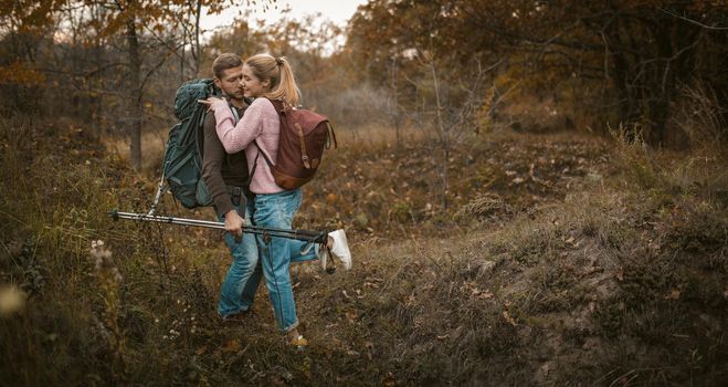 Couple of backpackers hiking in autumn forest outdoors, caucasian man and blonde woman traveling in nature with hiking poles. The guy helps his girlfriend supporting her on downhill.