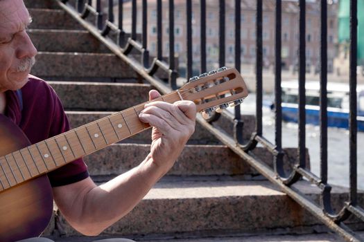 An adult elderly man of retirement age plays an old six-string classical acoustic guitar outdoors while sitting on a granite staircase on a summer evening in the city. Hobby. Selective focus.