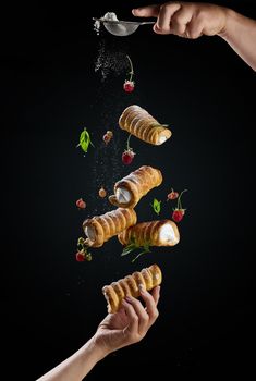 Baked crispy tubes with whipped protein cream levitate on a black background, powdered sugar pours from above