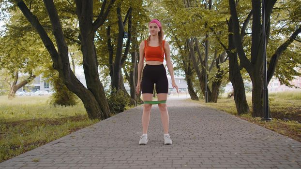Athletic fit slim sport girl doing squats using fitness elastic rubber band as part of her fitness workout outside in park at morning. Young woman enjoying healthy lifestyle. Active sportswoman jogger