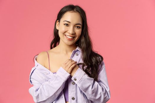 Close-up portrait of tender, romantic asian girl keep loving dearest memories in her heart, press hands to chest, smiling and looking thankful camera, standing pink background. Copy space