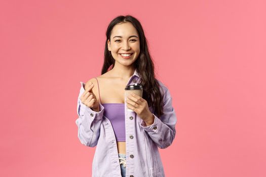 Happy stylish young modern female student walking from her favorite cafe, holding take-away coffee cup and smiling, drinking beverage enjoy sunny spring day, show love heart sign, pink background.