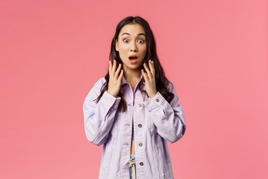 Astonished young korean girl seeing something shocking, stare camera with surprise, drop jaw and gasping, hold hands raised startled over big news, stand pink background.