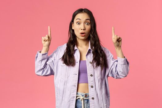 Portrait of impressed and surprised beautiful young girl in denim jacket, crop-top, pointing fingers up and staring at camera startled, gasping amazed, showing something cool, pink background.