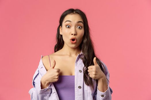 Close-up portrait of excited and amazed young asian woman, gasping seeing something stunning and awesome, show thumbs-up like, think product is super cool, stand pink background.