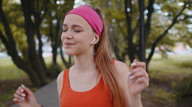 Athletic fitness sport runner girl training, listening favorite music song in earphones, looking at camera and smiling, laughing. Young woman working out cardio in park. Active sportswoman jogger