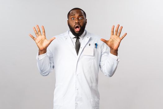 Portrait of shocked and nervous african-american male doctor raising hands in surrender, trying calm down tensed family of patient, look insecure and unaware what do, stand grey background.