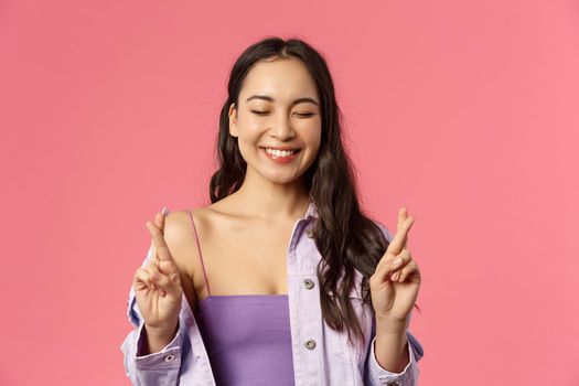 Close-up portrait of excited, optimistic and hopeful pretty girl in stylish outfit, cross fingers close eyes and smile as making wish, dreaming win competition, dream come true, pink background.