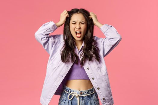 Portrait of furious young pissed-off girl losing her mind, distressed pull hair from head and screaming, grimacing look angry and hateful, standing stressed over pink background.